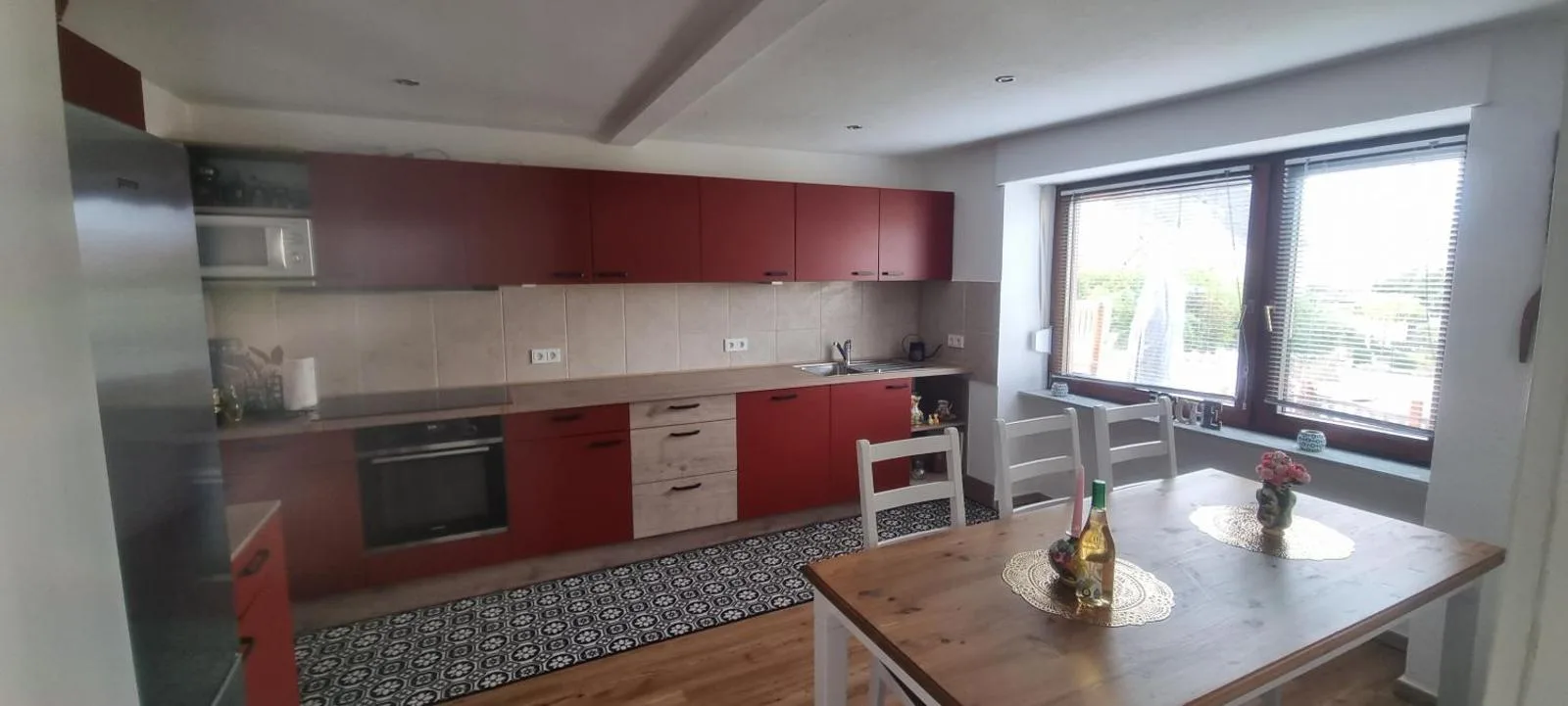 Große helle Küche mit roten Fronten, Edelstahlkühlschrank und Holzesstisch am großen Fenster mit viel Tageslicht.