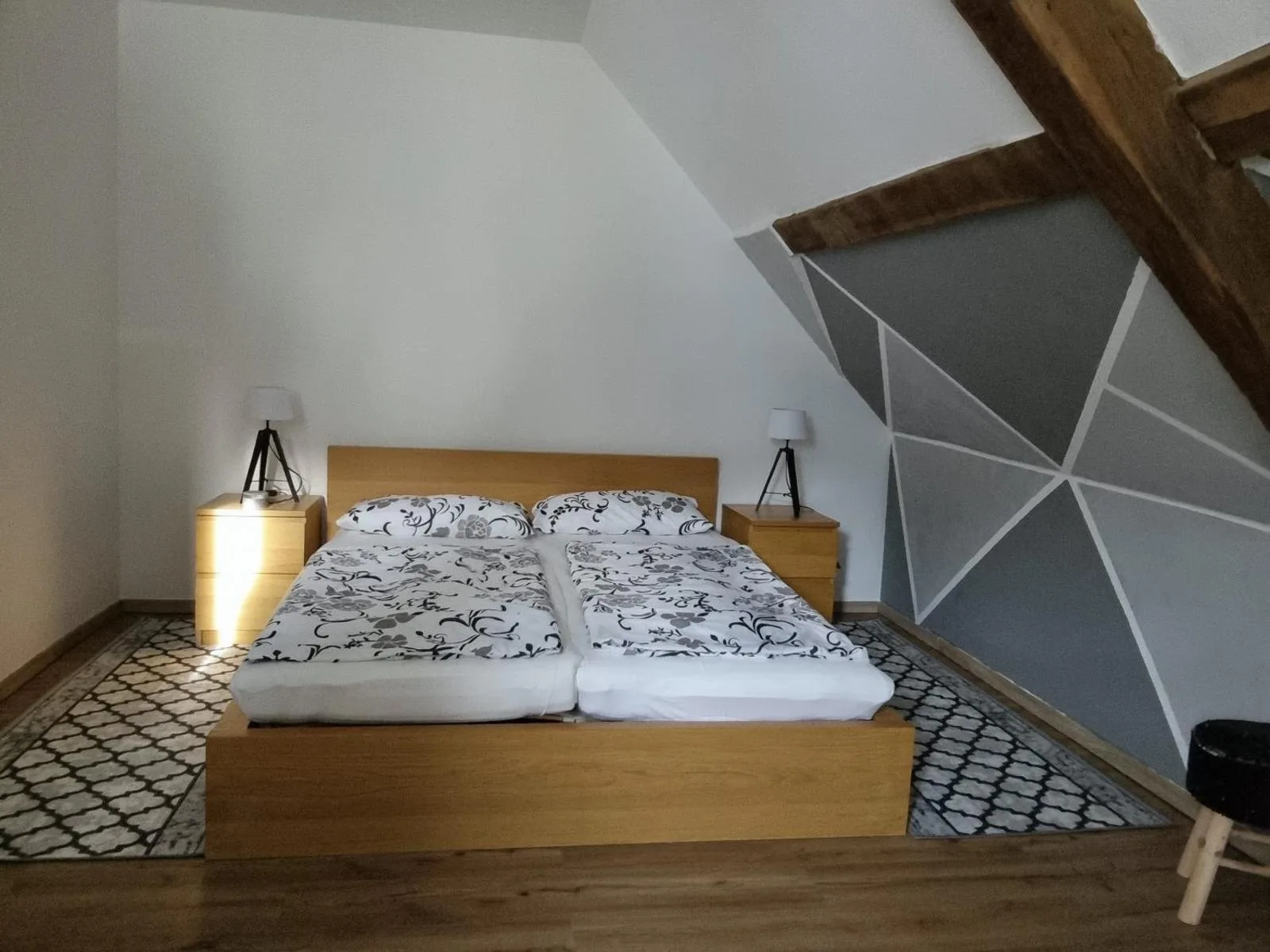 Doppelbett im Dachgeschoss unter sichtbaren Holzbalken vor grauer geometrischer Dreiecks-Wandgestaltung, gut ausgeleuchtet und aufgeräumt.