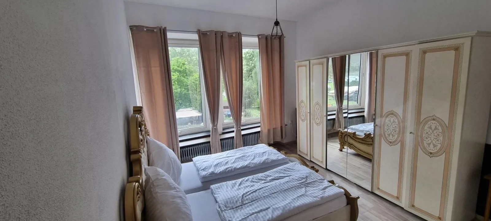 Schlafzimmer mit goldverziertem Doppelbett, spiegelbestücktem Kleiderschrank und rosafarbenen Vorhängen vor Fenster mit Grünblick