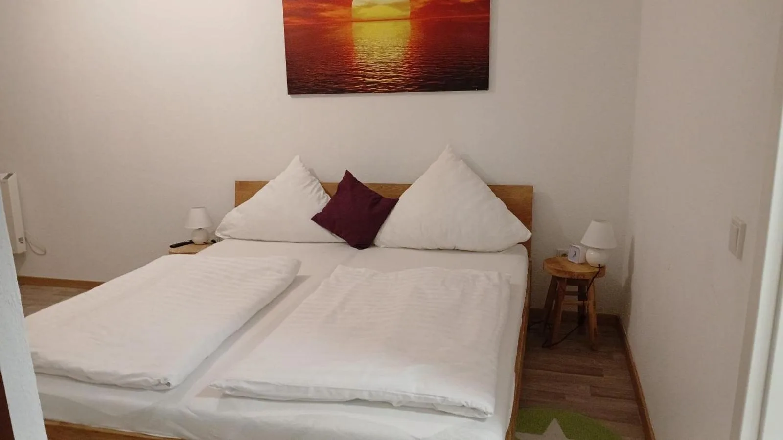 Doppelbett aus Holz mit weisser Bettwaesche und weinrotem Kissen, darueber ein Sonnenuntergangs-Bild ueber Wasser, Nachttisch mit Lampe.