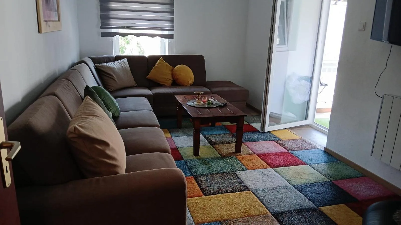 Wohnzimmer mit grosser brauner Ecksofa-Garnitur, bunten Kissen und einem farbenfrohen Patchwork-Teppich, Terrassentuer im Hintergrund offen.